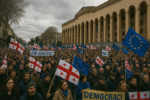 Wide protest on Rustaveli Avenue in Tbilisi, Georgia, with thousands of people waving Georgian and EU flags in front of the Parliament building under an overcast sky.