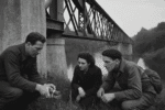 A black-and-white historical photograph showing three resistance fighters crouched near a railway bridge, examining an explosive device as they plan a sabotage operation during wartime.