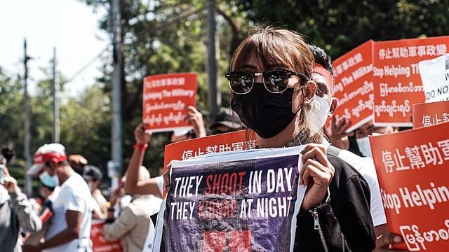 Masked protester in Myanmar holding a sign condemning military violence during anti-coup demonstrations, surrounded by others carrying red banners mobilizing resistance movements.