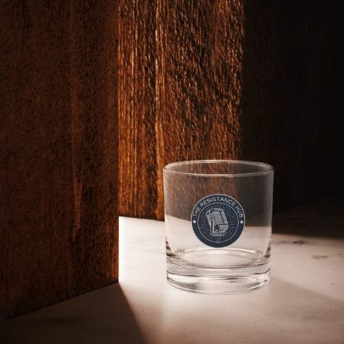 Clear lowball glass with navy Resistance Hub logo, placed on marble surface with wood panel shadows