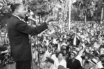 A civil rights leader speaks passionately at an outdoor rally, addressing a large crowd gathered in front of him. The audience, including men, women, and police officers, listens intently as he delivers his speech from a microphone on stage.