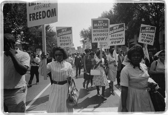 Civil rights demonstrators march along a roadway in Washington, D.C., holding signs calling for equal rights, integrated schools, fair housing, and an end to racial discrimination during the 1963 March on Washington.