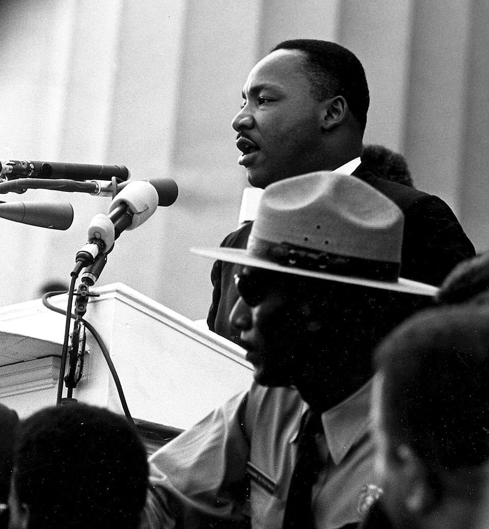 Dr. Martin Luther King Jr. speaks at a podium during the March on Washington in Washington, D.C., with microphones positioned in front of him and members of the crowd visible nearby, 1963.