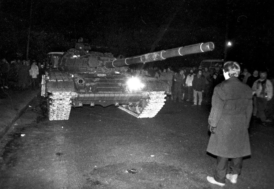 An unarmed Lithuanian citizen confronts a Soviet tank on a dark street in Vilnius during the January 13, 1991 assault on the television tower, with a crowd of civilian defenders visible in the background.