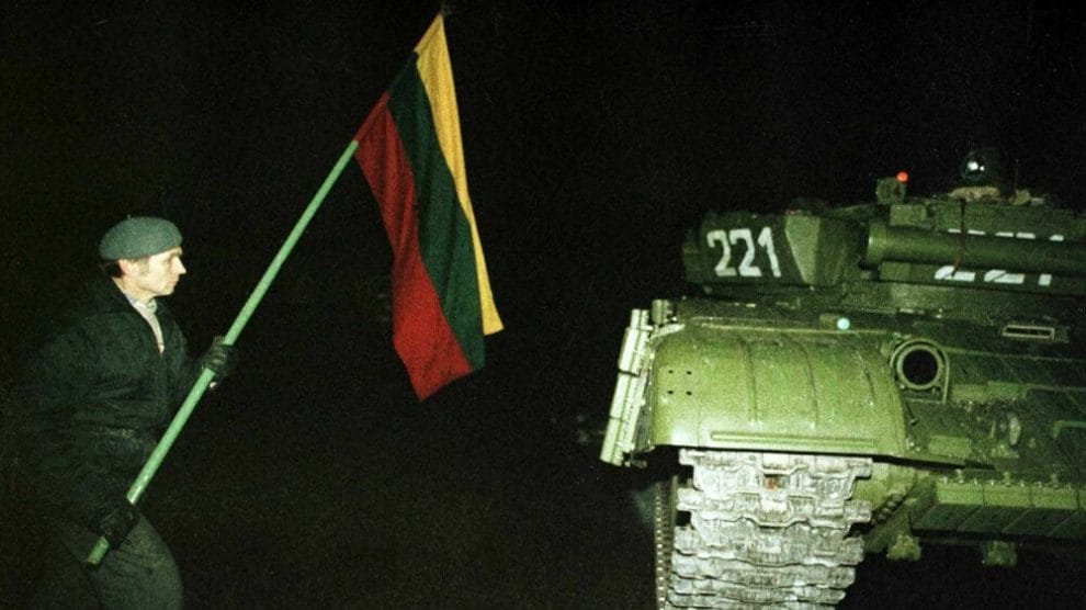 A man holding the Lithuanian national flag stands defiantly in front of a Soviet military tank at night, symbolizing resistance during the struggle for independence.
