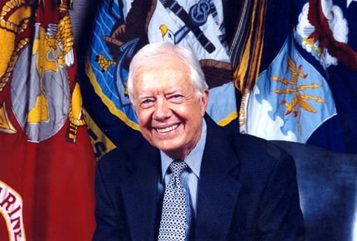 An elderly man in a suit and tie smiles while seated in front of U.S. military flags, including those of the Marine Corps, Navy, and Air Force.