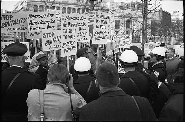 Civil rights demonstrators gather outside the White House holding signs demanding voting rights and protesting police brutality in Selma, Alabama, while law enforcement officers stand between protesters and the building, 1965.