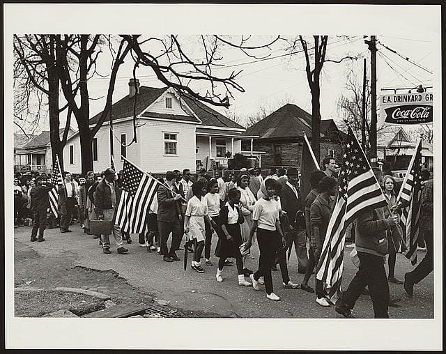 Civil rights marchers walk along a residential street during the Selma to Montgomery voting rights march in Alabama, carrying American flags and moving together in an organized procession, 1965.