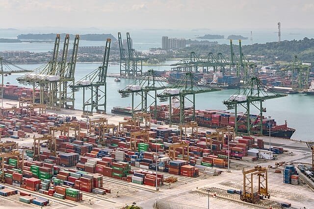 Wide view of the Port of Singapore showing container cranes, colorful cargo stacks, and docked ships along the waterfront, photographed from an elevated vantage point.