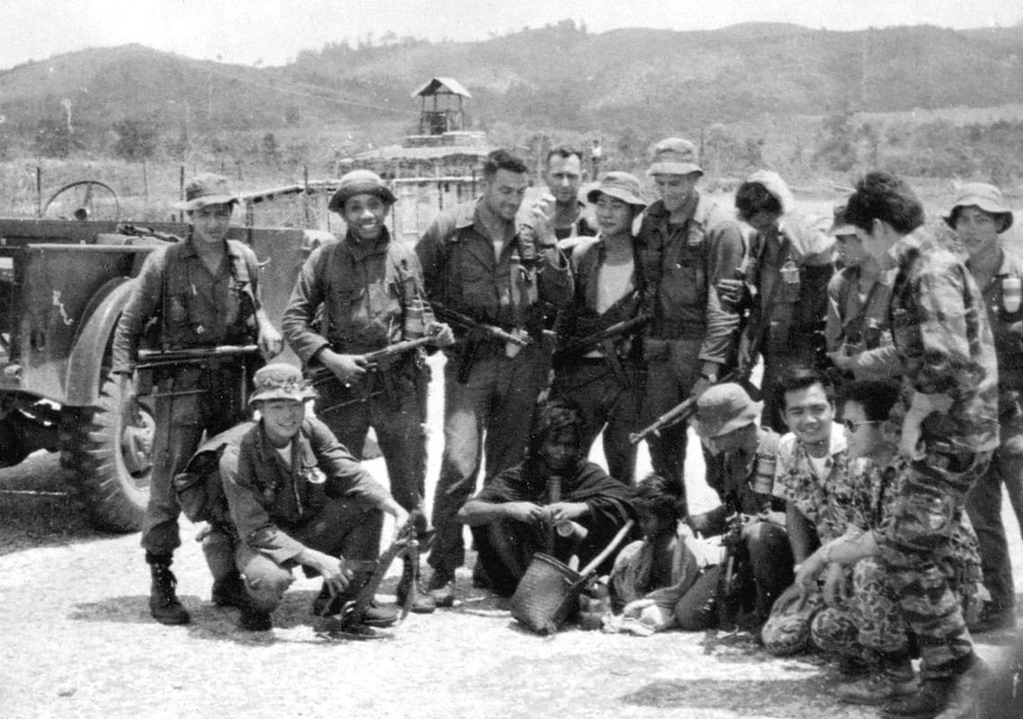 Black-and-white photograph of Major Richard Meadows (center, without hat) with a MACV-SOG reconnaissance team in Vietnam, 1966. The team poses with weapons and local personnel near a jeep and guard tower in the background. Cold War era evolution of elite warfare.