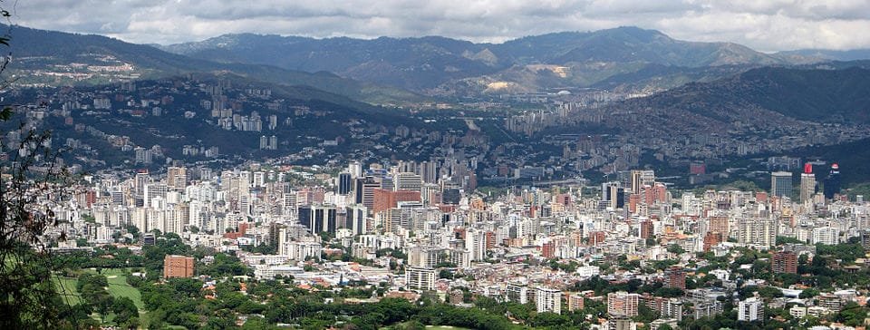 Panoramic view of Caracas showing dense high-rise development in the valley surrounded by steep, forested mountains, illustrating the city’s complex megacity terrain and compartmentalized urban structure.