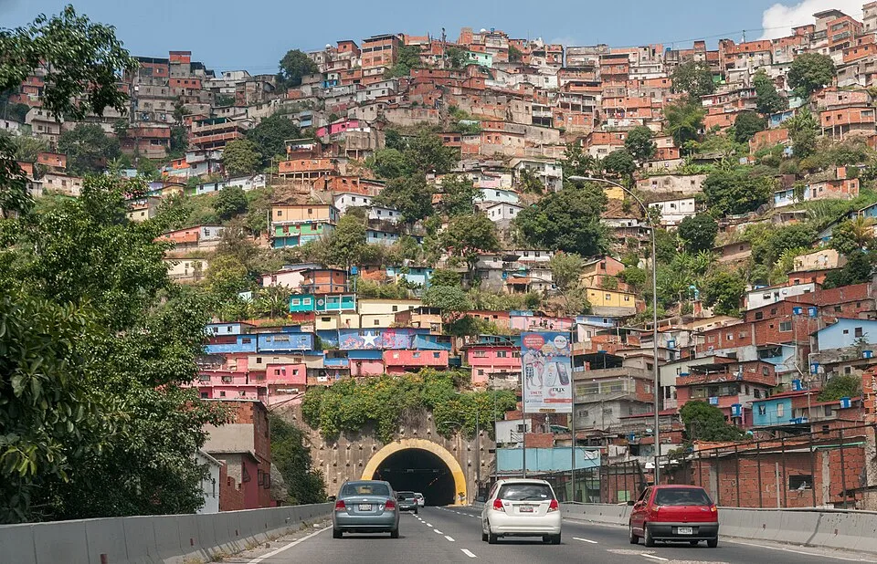 Hillside informal settlement in Caracas with dense clusters of brightly colored homes built on steep terrain above a highway tunnel.