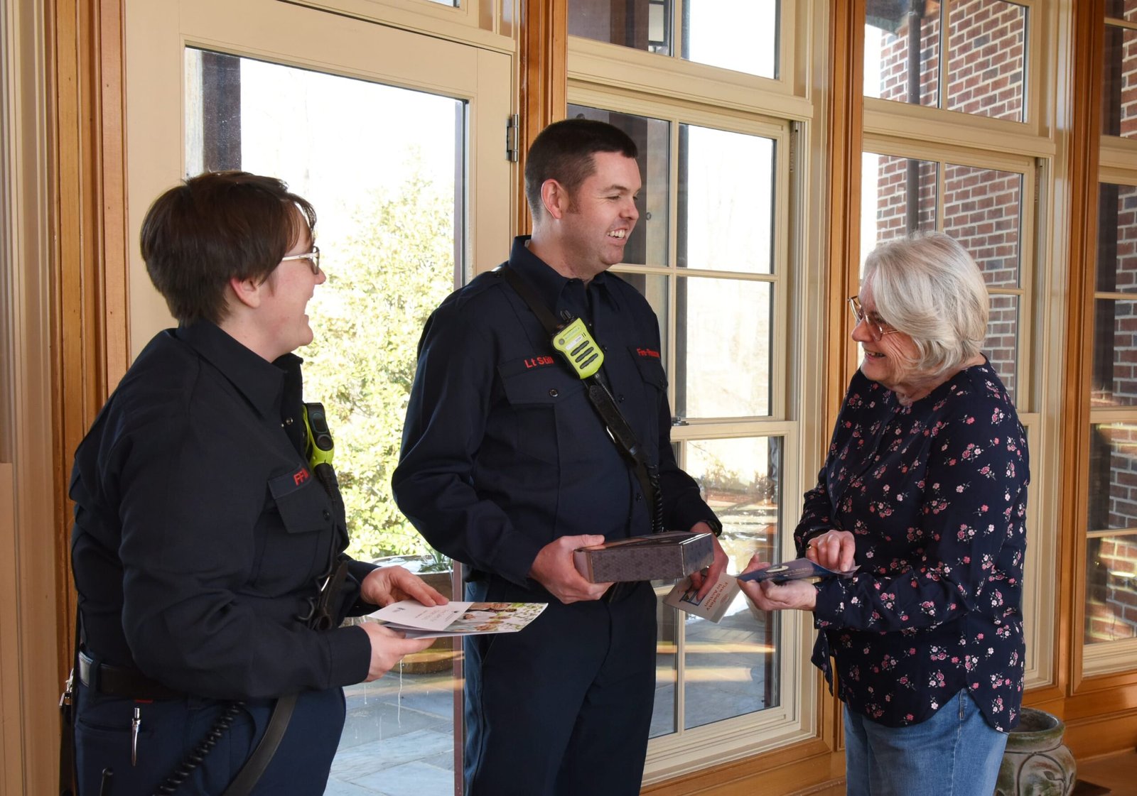 Two firefighters in uniform brief a community resident at her doorway during a wildfire preparedness or evacuation outreach visit.