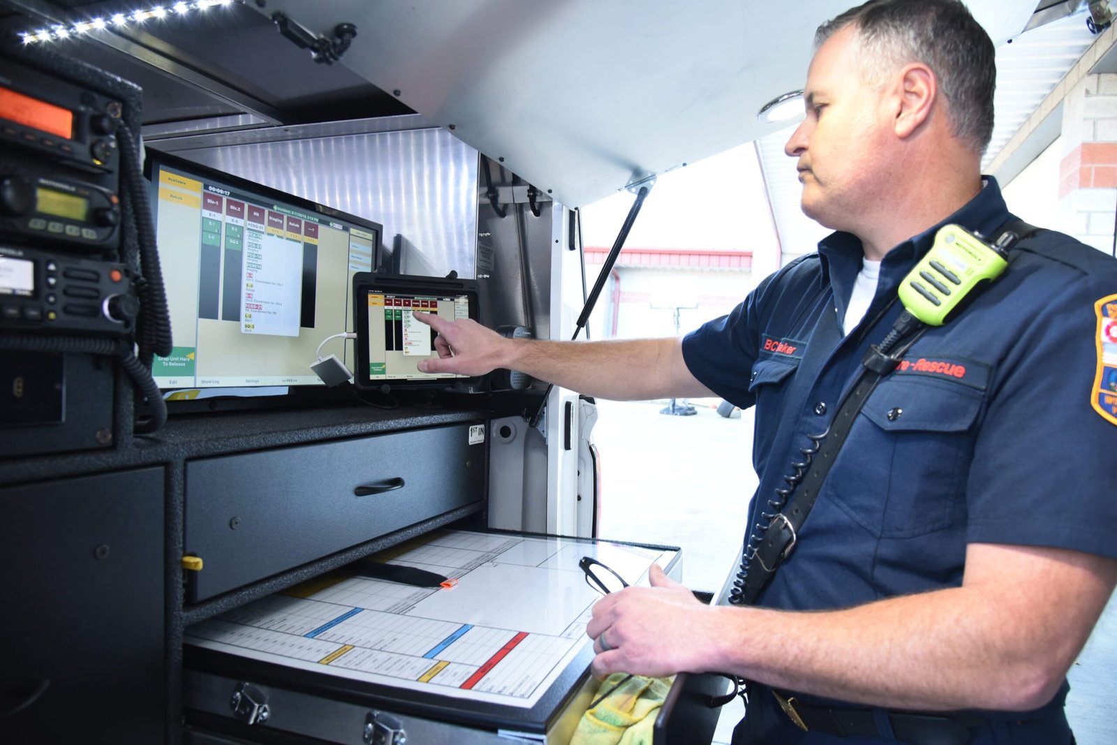 A fire officer monitors data on a laptop screen inside a mobile command vehicle, with radio equipment and operational documents visible.
