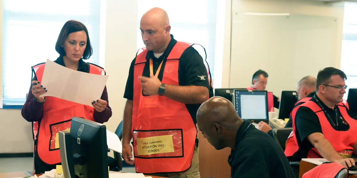 Emergency management personnel wearing orange Incident Command System vests coordinate operations inside a command center, with the Situation Unit Leader reviewing documents alongside team members at computer workstations.