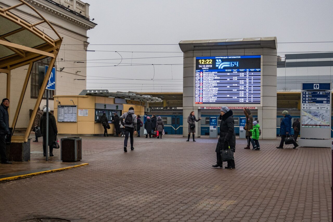Passengers at Minsk Passenger Railway Station in front of a digital Belarusian Railways departure board displaying train schedules.