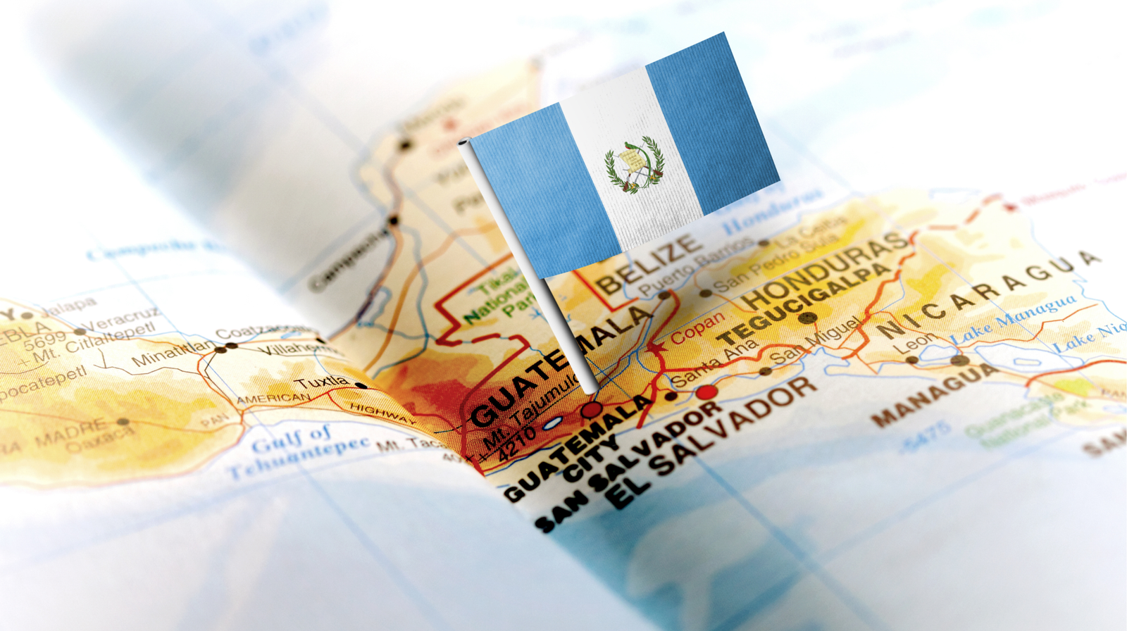 The flag of Guatemala on a small pin standing in a physical map of Central America, marking Guatemala City alongside neighboring Belize, Honduras, El Salvador, and Nicaragua.