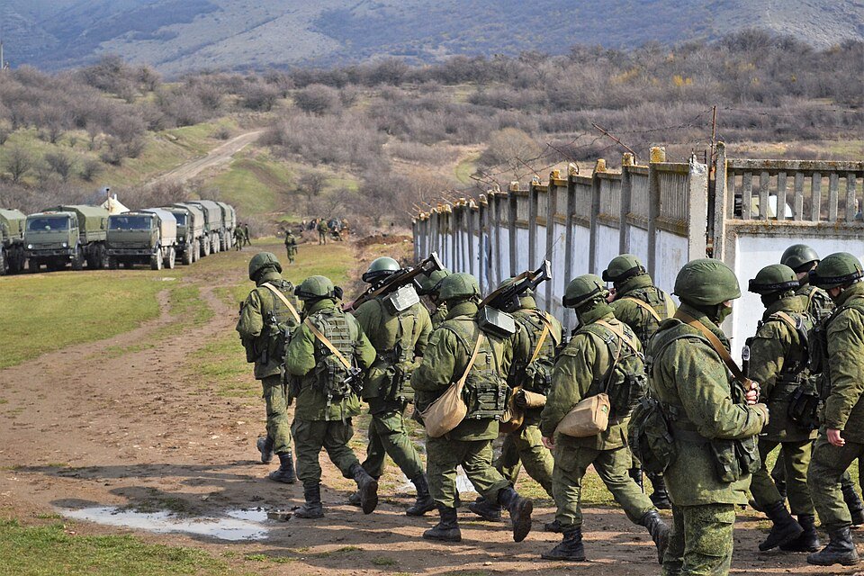 A column of Russian soldiers in green uniforms without insignia, carrying small arms and packs, march in formation toward the Perevalne military base in Crimea on 9 March 2014, with a line of military trucks visible behind a fence in the background, during the unmarked SPETSNAZ operation that became known as the Little Green Men deployment and that the ARIS study identifies as the mature form of Russian hybrid warfare