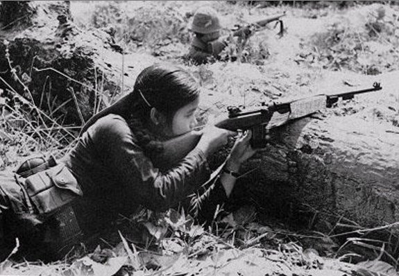 A female Viet Cong guerrilla lies prone in dense undergrowth aiming a rifle while a second fighter takes position behind her, illustrating the village-embedded irregular forces that formed the foundation layer of Vo Nguyen Giap's people's war system