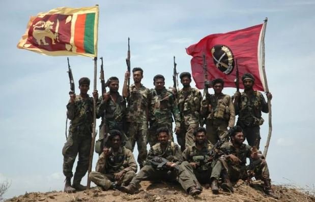 Sri Lankan Army soldiers pose on a rocky hilltop after the capture of Thoppigala, holding assault rifles and flying the national flag alongside a red unit colour.