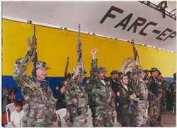 FARC-EP fighters in camouflage raising their rifles in front of a wall painted with the guerrilla group’s name and the Colombian flag colors