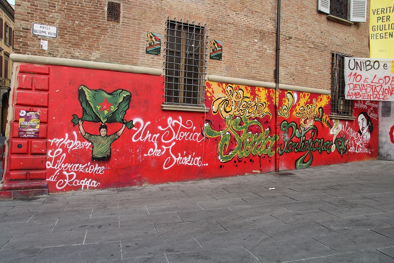 Street mural on a red-painted wall in Piazza Verdi, Bologna, showing a figure in a green field uniform holding the YPJ flag overhead beside handwritten Italian text commemorating the liberation of Raqqa on 17 October 2017, with the slogan Storie d'oggi, Storia Partigiana painted in green and gold across the rest of the wall
